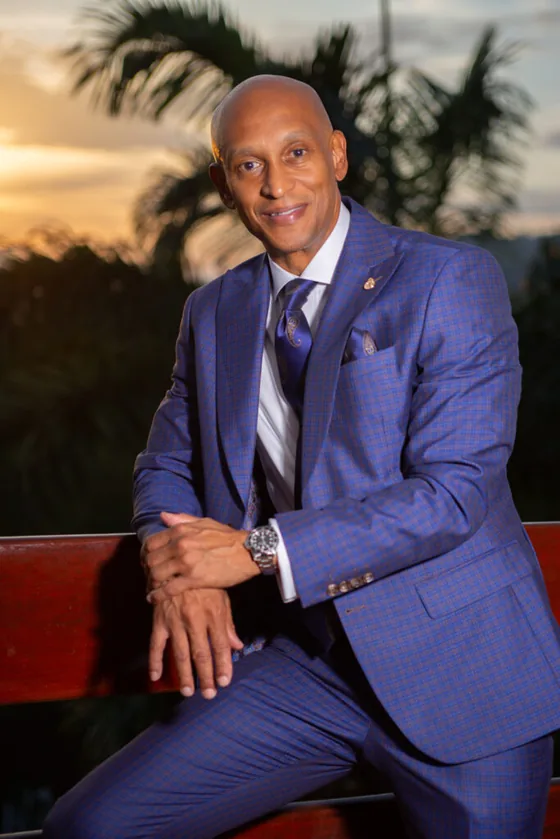 Gregory Arneaud in a blue checked suit, seated outdoors at sunset with palm trees behind him